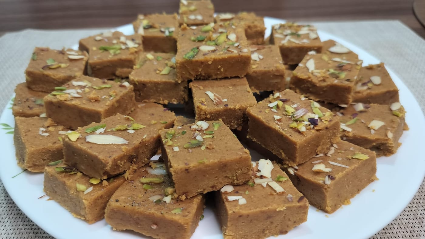 Squares of golden besan barfi topped with slivered almonds and pistachio on a white plate