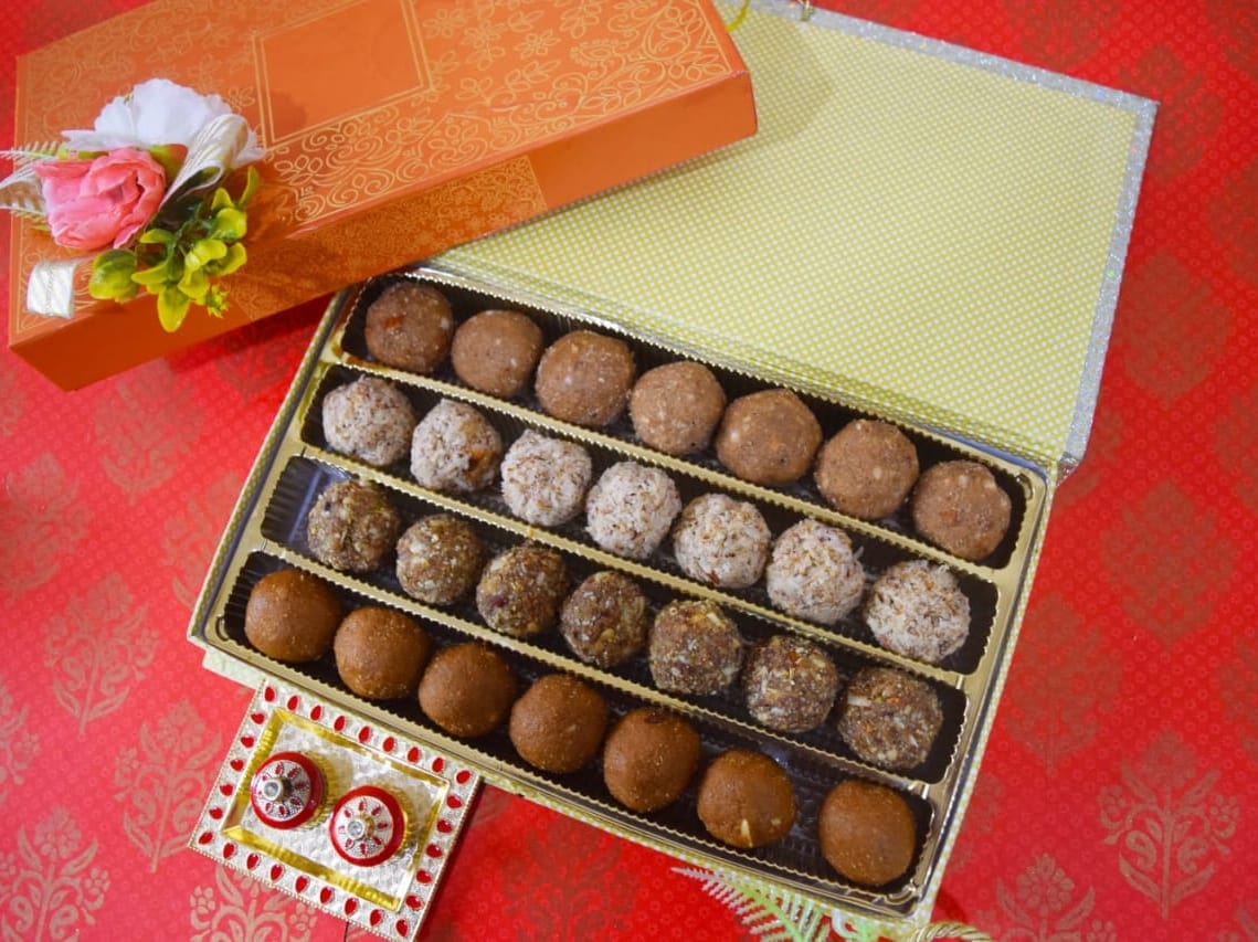 An open gift box with four rows of assorted homemade millet laddoos, styled for a festive occasion